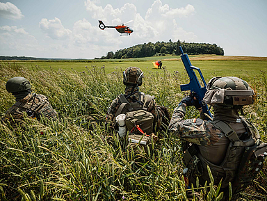 Drei Soldaten in Tarnuniform liegen im hohen Gras, einer hält ein blaues Übungsgewehr. Im Hintergrund fliegt ein orangefarbener Hubschrauber über ein Feld unter blauem Himmel. Das Bild zeigt drei Soldaten in Tarnuniform und Helmen, die im hohen Gras eines Feldes liegen. Einer trägt ein blaues Übungsgewehr. Im Hintergrund ist ein orangefarbener Hubschrauber in der Luft zu sehen, dahinter eine Baumreihe und ein weiter Himmel mit Wolken.