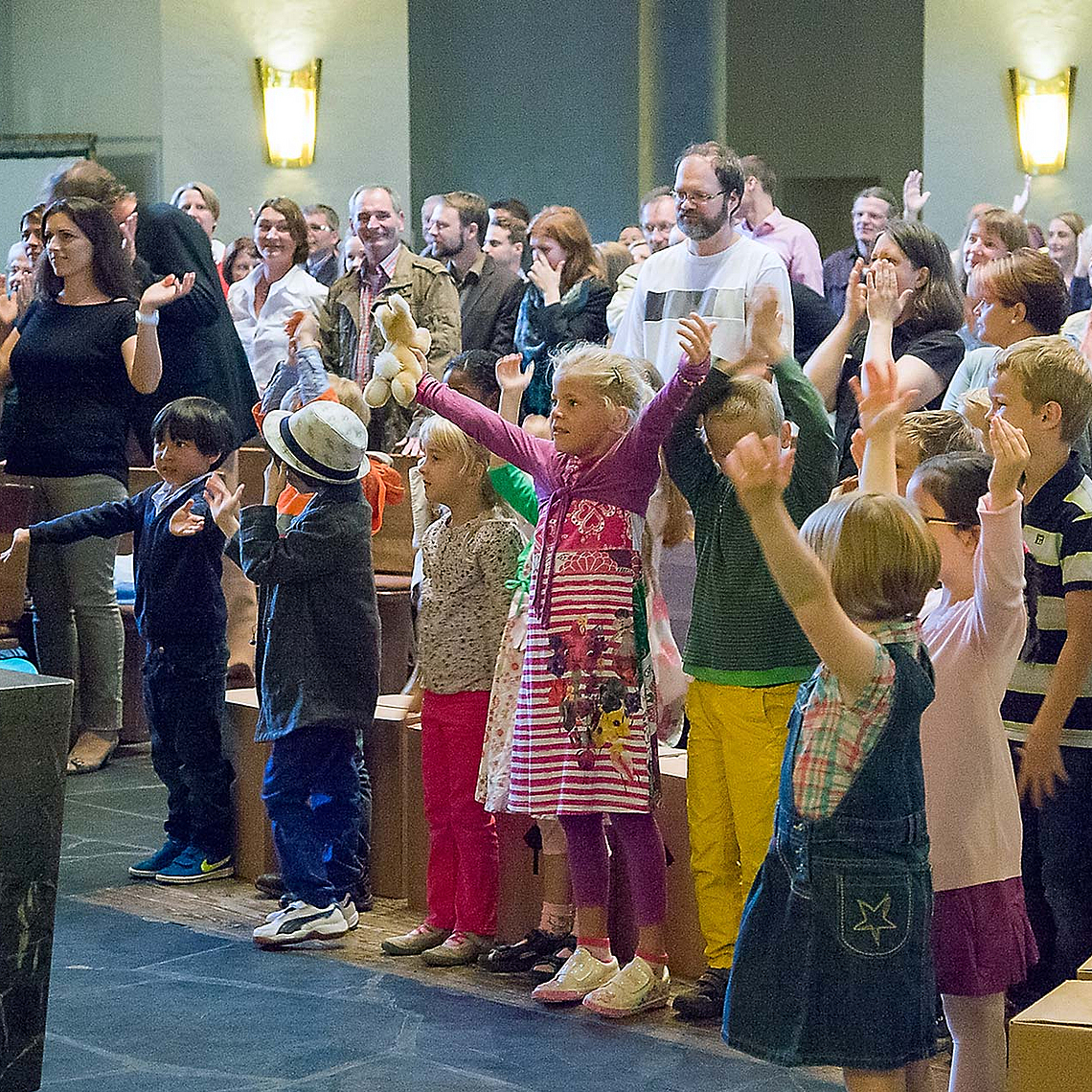 Kinder stehen mit erhobenen Armen in der ersten Reihe eines gut besuchten Gottesdienstes, Erwachsene sitzen und stehen dahinter in einem hellen Kirchenraum mit Holzbänken und hoher Decke. Auf dem Boden liegen Kartons. Kinder stehen mit erhobenen Armen in der ersten Reihe eines gut besuchten Gottesdienstes, Erwachsene sitzen und stehen dahinter in einem hellen Kirchenraum mit Holzbänken und hoher Decke. Auf dem Boden liegen Kartons.