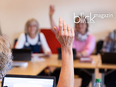 Drei Personen sitzen an einem Tisch mit Papieren und Stiften, im Hintergrund ein Bücherregal. Die Personen sind in ein Gespräch vertieft. Die Szene spielt in einem hellen Raum mit Tageslicht. Das Foto zeigt drei Personen, die nebeneinander an einem Tisch sitzen. Auf dem Tisch liegen mehrere Blätter Papier, ein Notizbuch und Stifte. Die Personen sind in ein Gespräch vertieft und blicken auf die Unterlagen. Im Hintergrund steht ein großes Bücherregal, das mit zahlreichen Büchern gefüllt ist. Der Raum ist hell und freundlich, Tageslicht fällt durch ein Fenster ein. Die Szene vermittelt eine konzentrierte und kooperative Arbeitsatmosphäre.