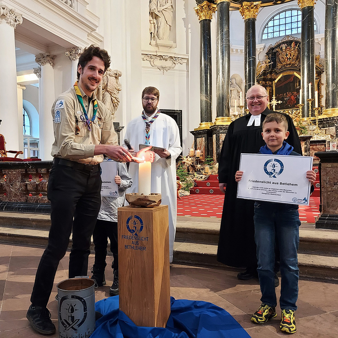 Friedenslicht-Träger 2025 Fabian Stork (DPSG, Stamm Wächtersbach) entzündet eine Kerze mit dem Friedenslicht aus Bethlehem. Friedenslicht-Träger 2025 Fabian Stork (DPSG, Stamm Wächtersbach) entzündet eine Kerze mit dem Friedenslicht aus Bethlehem. Im Hintergrund zu sehen sind DPSG-Diözesankurat Patrick Jestädt (l.) und Pfarrer Jörg Scheer von der evangelischen Christuskirche in Fulda.
