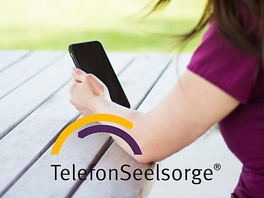 Person hält ein Smartphone in der Hand. Das Logo der TelefonSeelsorge ist in das Bild eingeblendet. Eine Person sitzt an einem Holztisch im Freien und hält ein Smartphone in der Hand. Im Bild ist das Logo der TelefonSeelsorge zu sehen. Das Foto steht symbolisch für die telefonische und digitale Beratung von Menschen in Krisen.