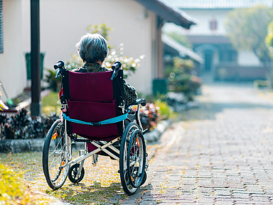 Person in einem Rollstuhl auf einem gepflasterten Weg, im Hintergrund Häuser und Grün. Eine Person sitzt in einem Rollstuhl und ist von hinten zu sehen. Der Rollstuhl steht auf einem gepflasterten Weg, der leicht nach vorne verläuft. Links und rechts des Weges sind Pflanzen und Sträucher zu erkennen. Im Hintergrund befinden sich Häuser mit hellen Fassaden und ein Dach mit grünen Ziegeln. Die Szene wirkt ruhig und zeigt eine Außenumgebung bei Tageslicht.