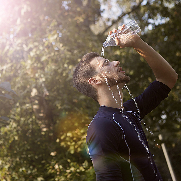 Person kühlt sich an einem heißen Sommertag mit Wasser aus einer Trinkflasche. Das Bild veranschaulicht die Auswirkungen extremer Hitze. Eine Person gießt sich im Freien Wasser aus einer Plastikflasche über Gesicht und Kopf. Im Hintergrund sind Bäume und helles Sonnenlicht zu sehen. Das Motiv steht symbolisch für hohe Temperaturen und die Belastungen durch Hitzewellen.