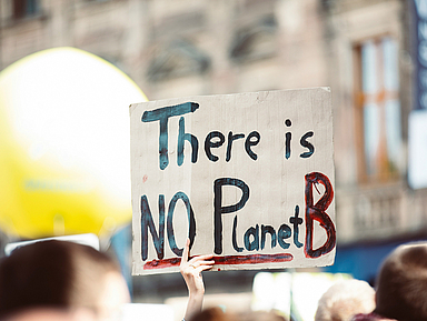 Hand hält ein Schild mit der Aufschrift „There is NO Planet B“ bei einer Demonstration im Freien, unscharfer Hintergrund mit Menschen und Gebäuden. Bei einer Demonstration im Freien hält eine Person ein handgemaltes Schild hoch. Darauf steht in großen Buchstaben: „There is NO Planet B“. Im Hintergrund sind weitere Menschen, ein großes gelbes Objekt und unscharfe Gebäude zu sehen.