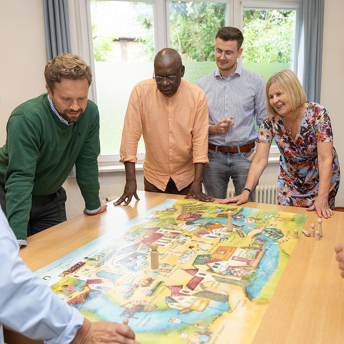 Planspiel zur Kirchenvorstandsarbeit mit acht Personen um einen Tisch, darunter Pfarrerin Ulrike Joachimi (4.v.r.), Beauftragte für die Kirchenvorstandswahl 2025 in der EKKW. Sieben Personen stehen oder lehnen sich um einen Tisch, auf dem ein großes, buntes Spielbrett liegt. Es handelt sich um ein Planspiel zur Kirchenvorstandsarbeit. Die Gruppe wirkt konzentriert und diskutiert offenbar aktiv über das Spielgeschehen. Pfarrerin Ulrike Joachimi (vierte von rechts) nimmt ebenfalls teil. Im Hintergrund ist ein Fenster mit Blick ins Grüne zu sehen.
