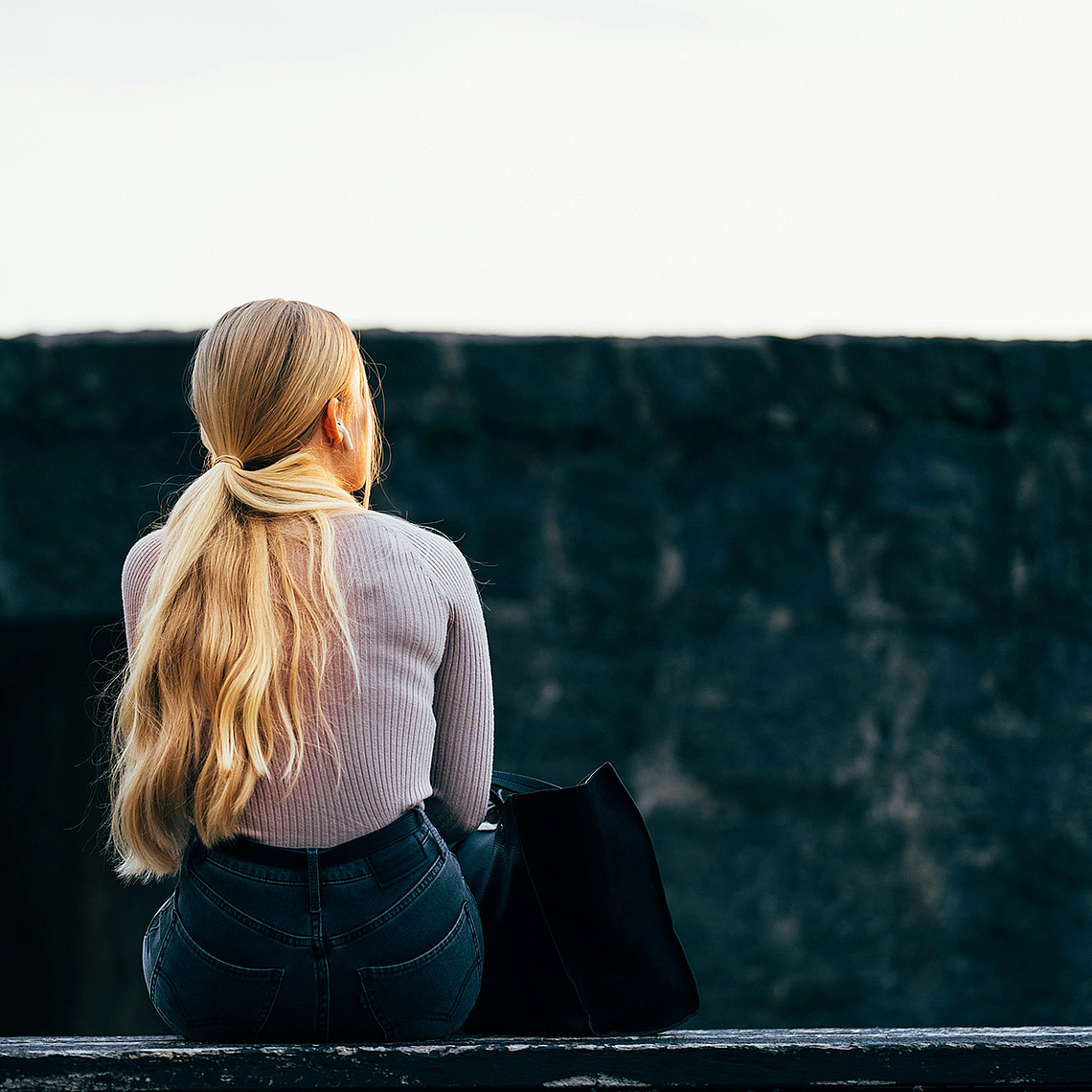 Das Foto zeigt eine Frau von hinten, die alleine auf einer Mauer sitzt. Das Foto zeigt eine Frau von hinten, die alleine auf einer Mauer sitzt.