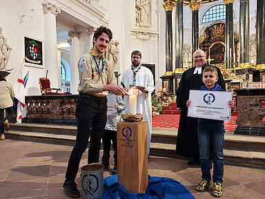 Friedenslicht-Träger 2025 Fabian Stork (DPSG, Stamm Wächtersbach) entzündet eine Kerze mit dem Friedenslicht aus Bethlehem. Friedenslicht-Träger 2025 Fabian Stork (DPSG, Stamm Wächtersbach) entzündet eine Kerze mit dem Friedenslicht aus Bethlehem. Im Hintergrund zu sehen sind DPSG-Diözesankurat Patrick Jestädt (l.) und Pfarrer Jörg Scheer von der evangelischen Christuskirche in Fulda.
