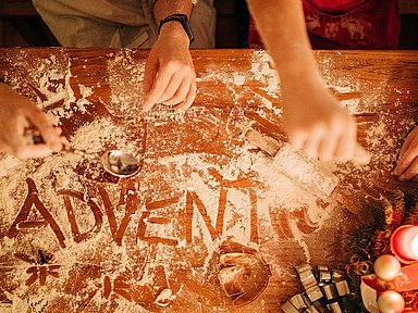  Zwei Personen backen Plätzchen. In das Mehl auf dem Tisch ist das Wort Advent geschrieben.