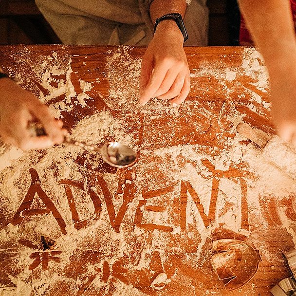  Zwei Personen backen Plätzchen. In das Mehl auf dem Tisch ist das Wort Advent geschrieben.