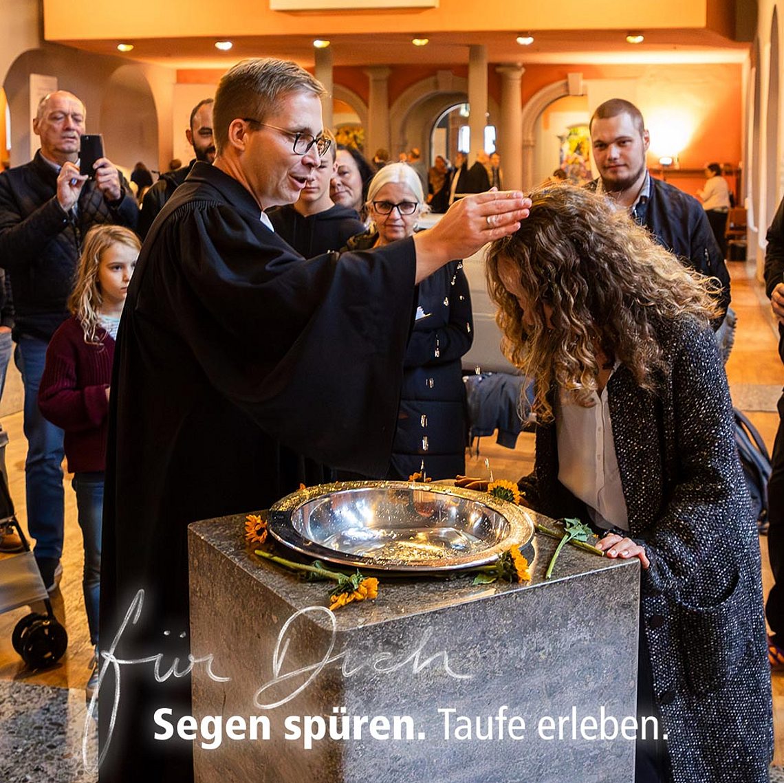 Taufe in der Kirche mit individueller Gestaltung – eine Frau wird am Taufbecken getauft. Eine Frau wird in einer evangelischen Kirche am Taufbecken von einem Pfarrer mit Wasser getauft. Um sie herum stehen Erwachsene und Kinder. Im Hintergrund sind Kirchenbänke, eine Orgel und ein Kinderwagen zu sehen. Text im Bild: ‚Für Dich. Segen spüren. Taufe erleben.