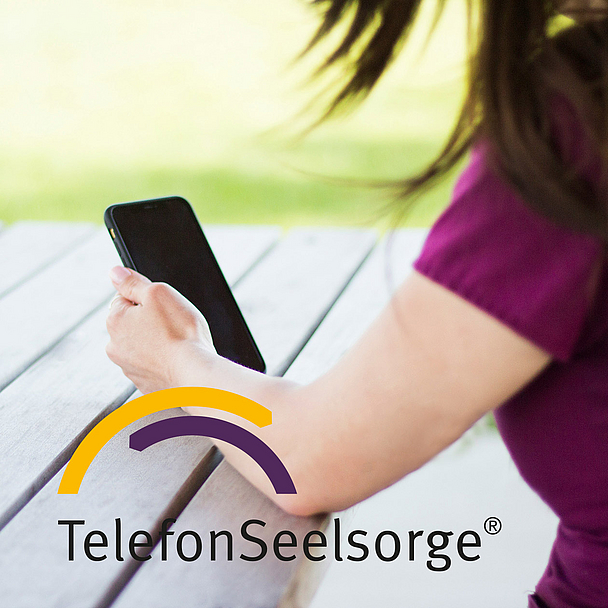 Person hält ein Smartphone in der Hand. Das Logo der TelefonSeelsorge ist in das Bild eingeblendet. Eine Person sitzt an einem Holztisch im Freien und hält ein Smartphone in der Hand. Im Bild ist das Logo der TelefonSeelsorge zu sehen. Das Foto steht symbolisch für die telefonische und digitale Beratung von Menschen in Krisen.
