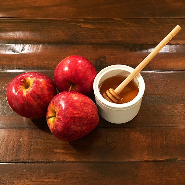 Äpfel und Honig stehen zu Rosch Haschana traditionell für die Hoffnung auf ein gutes und süßes neues Jahr. Drei rote Äpfel liegen neben einer Schale mit Honig und einem Honiglöffel auf einem dunklen Holztisch. Äpfel mit Honig gehören zu den traditionellen Speisen des jüdischen Neujahrsfestes Rosch Haschana.