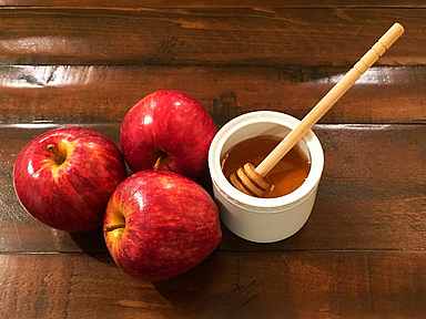 Äpfel und Honig stehen zu Rosch Haschana traditionell für die Hoffnung auf ein gutes und süßes neues Jahr. Drei rote Äpfel liegen neben einer Schale mit Honig und einem Honiglöffel auf einem dunklen Holztisch. Äpfel mit Honig gehören zu den traditionellen Speisen des jüdischen Neujahrsfestes Rosch Haschana.