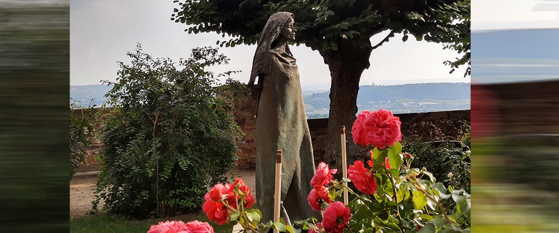 Darstellung der Hildegard von Bingen als Bronzeskulptur von Karlheinz Oswald vor Kloster Eibingen in einem Garten mit roten Rose und Bäumen, in der Ferne die Rochuskapelle von Bingen. (Foto: Wikipedia/Gerda Arendt)